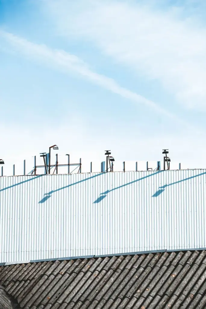 Corrugated metal wall with mounted light fixtures casting shadows above a tiled roof edge under blue sky