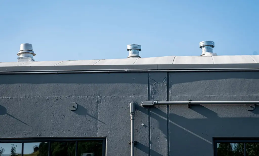 Three metal vent caps along the edge of a gray commercial building roof with conduit and windows below