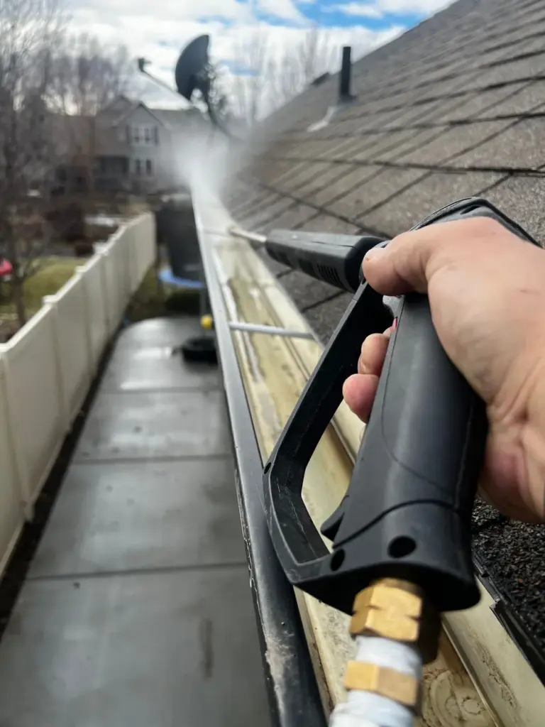Hand holding pressure washer wand spraying debris from a roof gutter on a shingle roof