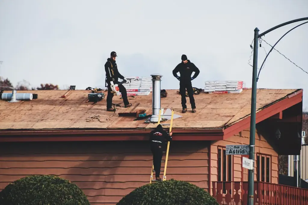 Three roofers on a bare wood roof deck with shingle bundles, one climbing a ladder, Cypress, TX