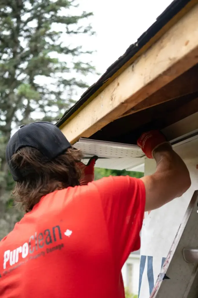 Worker in red shirt installing a soffit panel under a roof eave with exposed wood decking